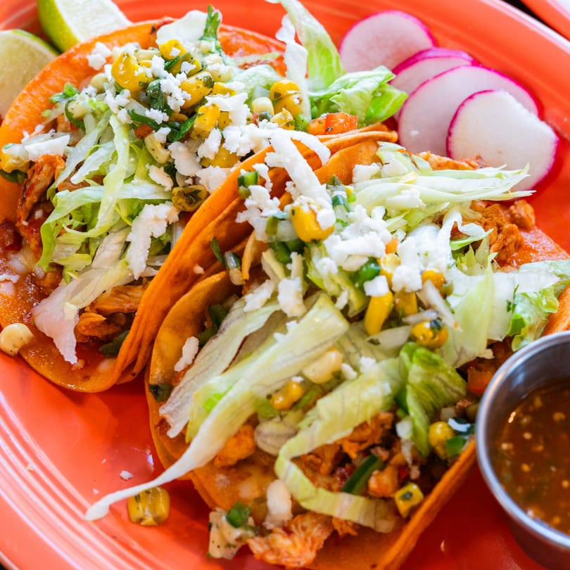 Three vibrant orange tacos topped with lettuce, corn, and cheese, served with radishes and lime.