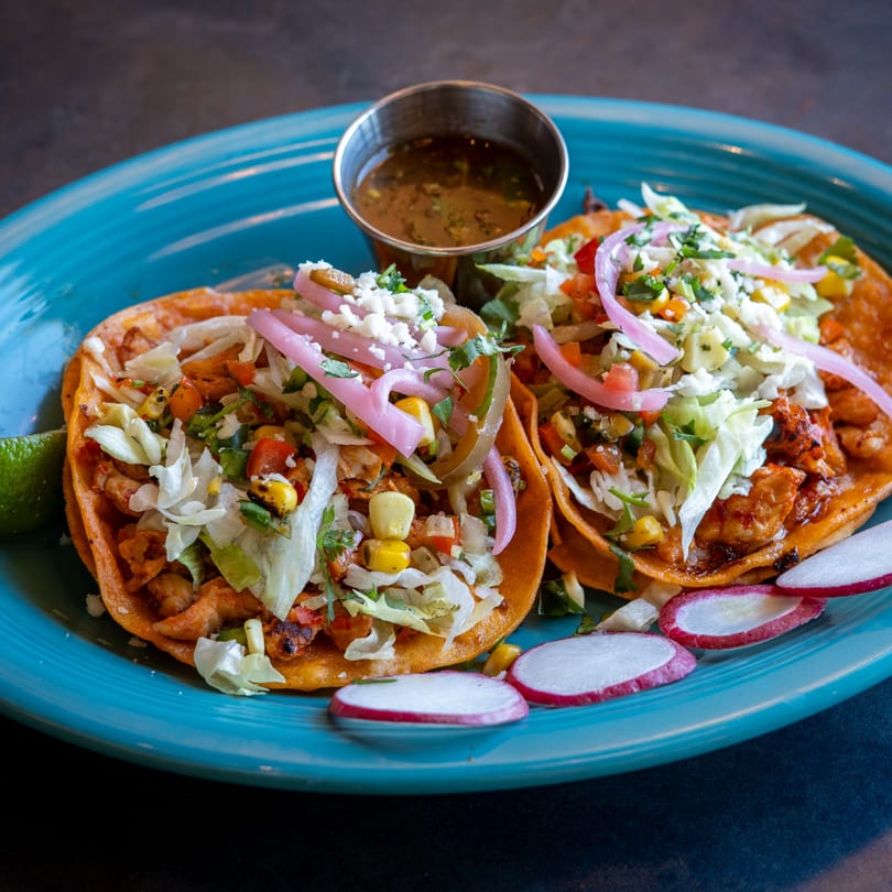 Two vibrant shrimp tacos with pickled onions, corn, and cheese, served with a side of sauce.