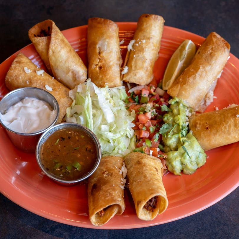 A vibrant orange plate filled with taquitos, guacamole, pico de gallo, shredded lettuce, and dipping sauces.
