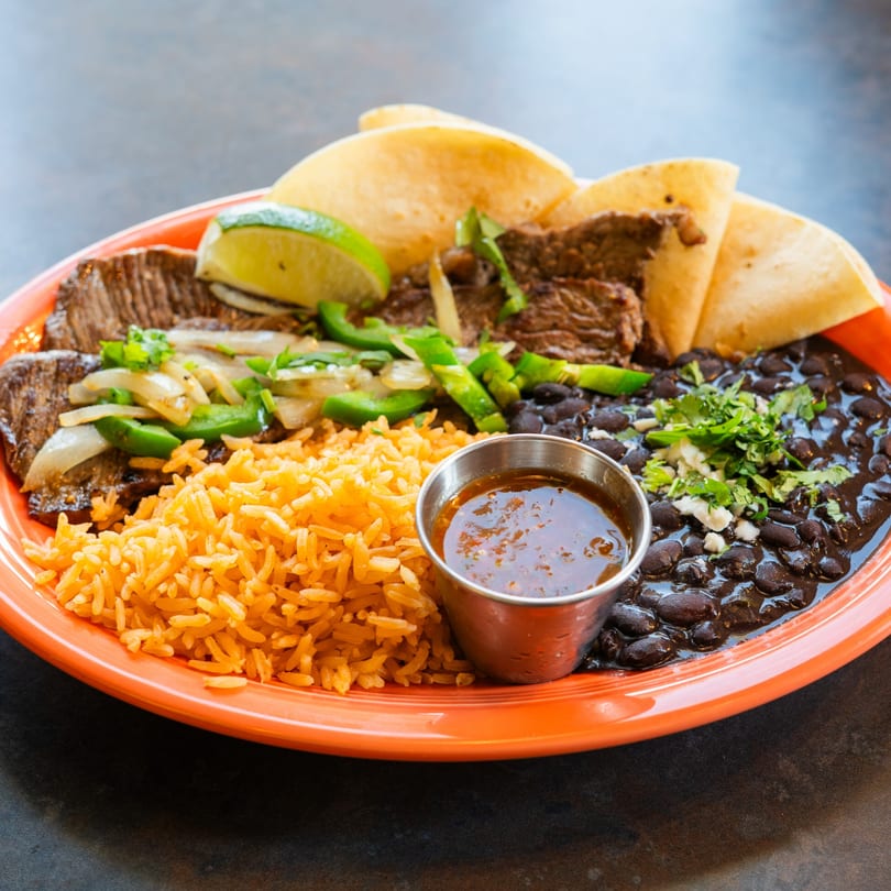 Steak fajitas with grilled onions, peppers, rice, black beans, tortillas, and a side of sauce.