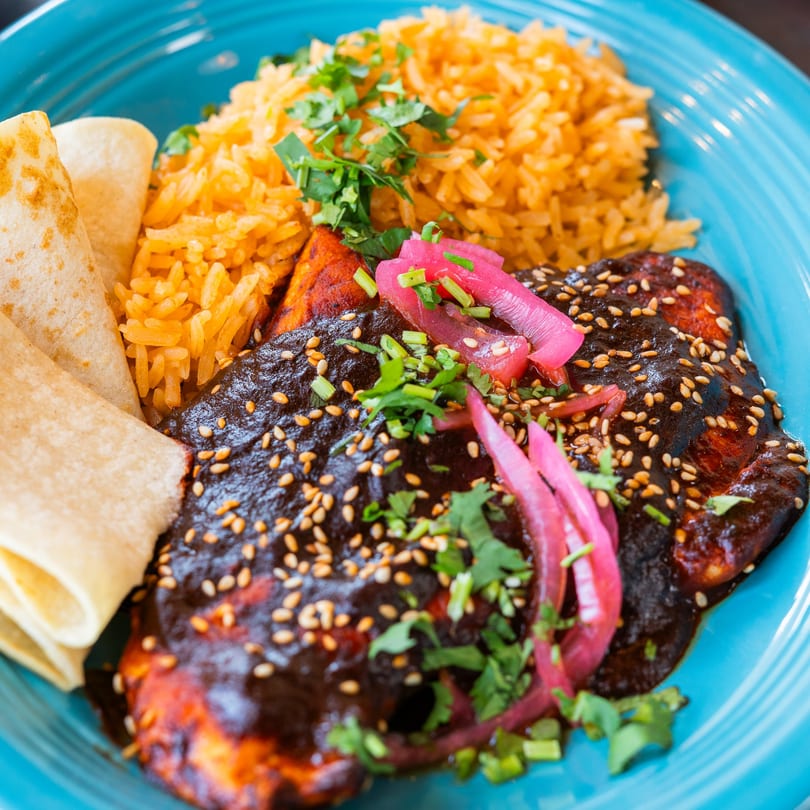 Chicken mole with rice, tortillas, pickled onions, and sesame seeds on a blue plate.