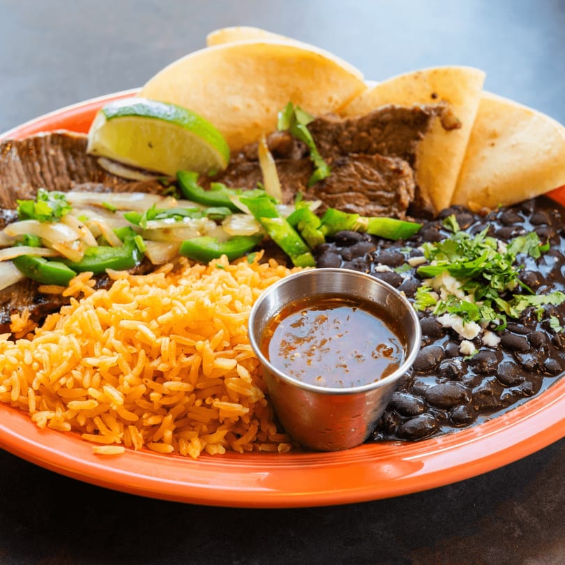 Carne asada with rice, black beans, tortillas, lime, and salsa on an orange plate.
